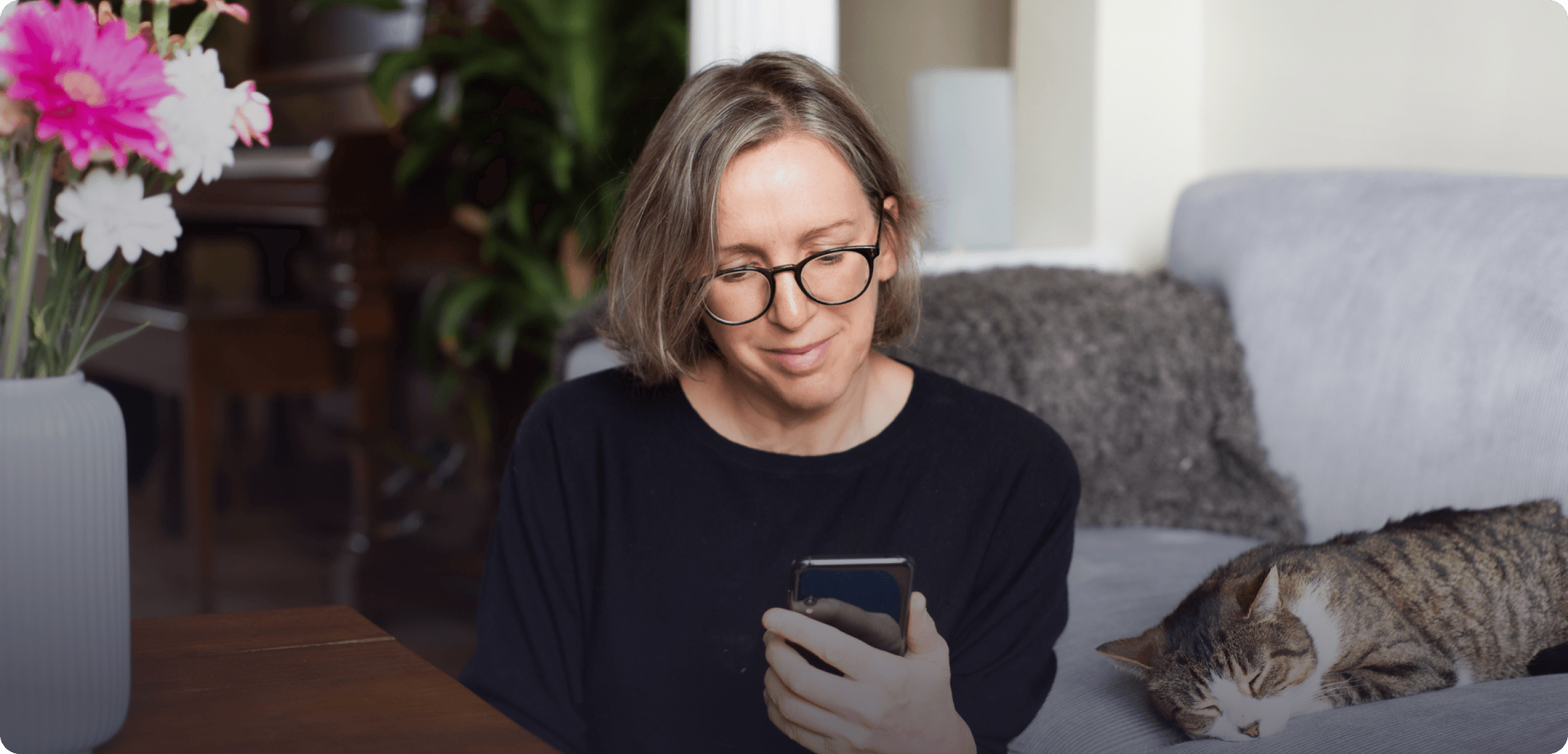 A woman sitting on the floor of her living room, looking at her phone while her cat lays next to her on the couch