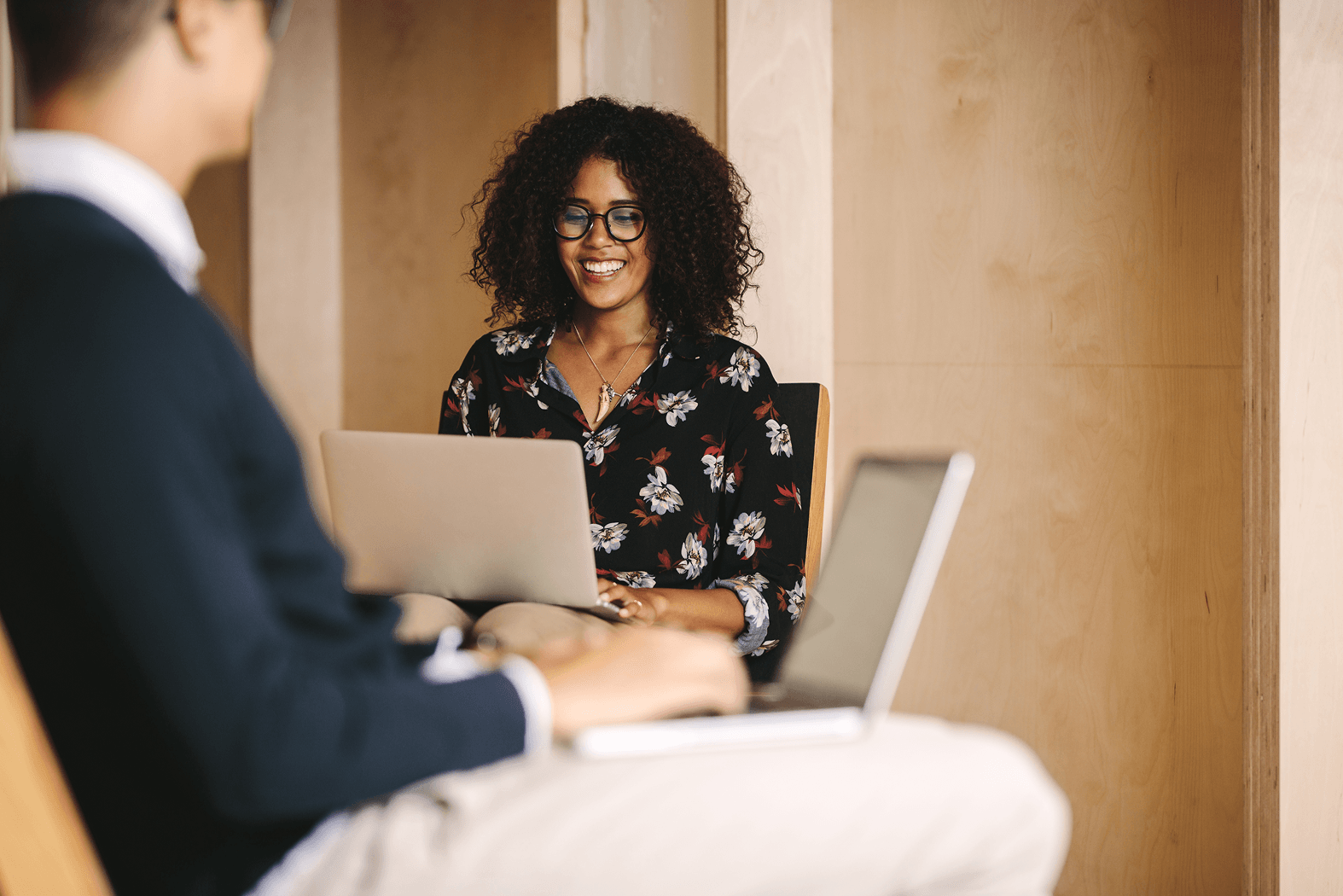 A woman with curly hair and glasses smiles while working on a laptop in a cozy, wooden environment. A man is partially visible in the foreground, also using a laptop.